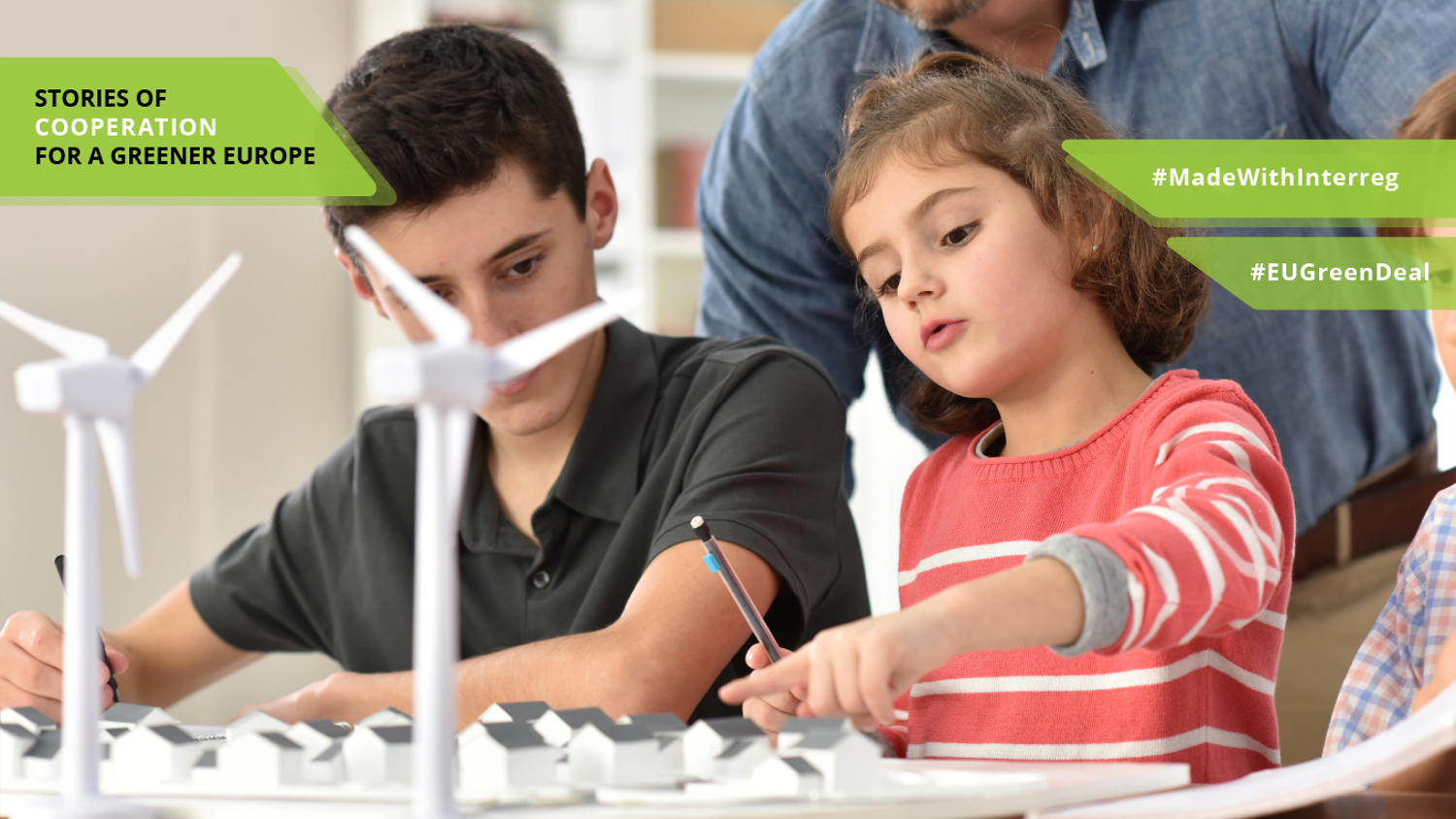 A girl and a boy work on a wind turbine model. An adult observes and assists. The image promotes environmental education with captions: "Stories of Cooperation for a Greener Europe," "#MadeWithInterreg," and "#EUGreenDeal.
