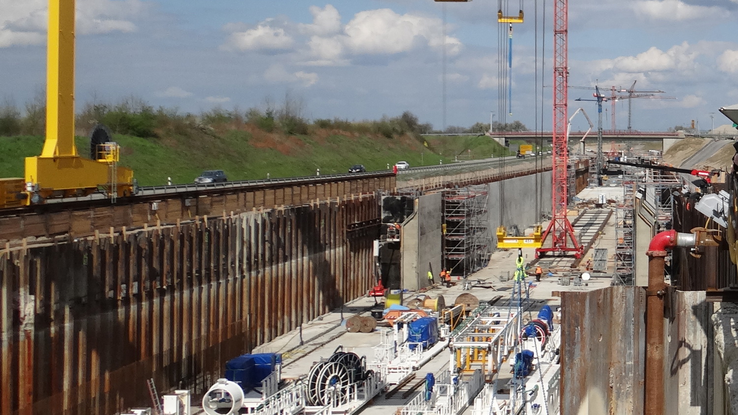 Construction site with large yellow cranes above a channel. Machinery and large pipes are visible along the ground. Workers can be seen in the distance. Earth and clouds in the background.