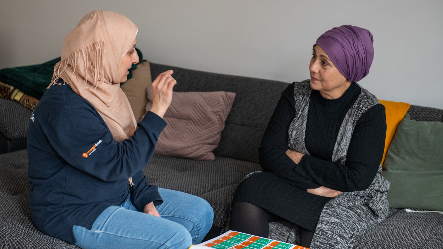 Two women in headscarves sit on a couch having a conversation. One gestures while speaking, and the other listens with arms crossed. Papers and a colorful chart are on the table in front of them.