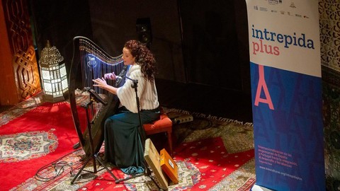 A woman sits on a red stool playing a harp on a patterned carpeted stage next to a lantern. A blue and white "intrepida plus" banner stands nearby, with ornate decor in the background.