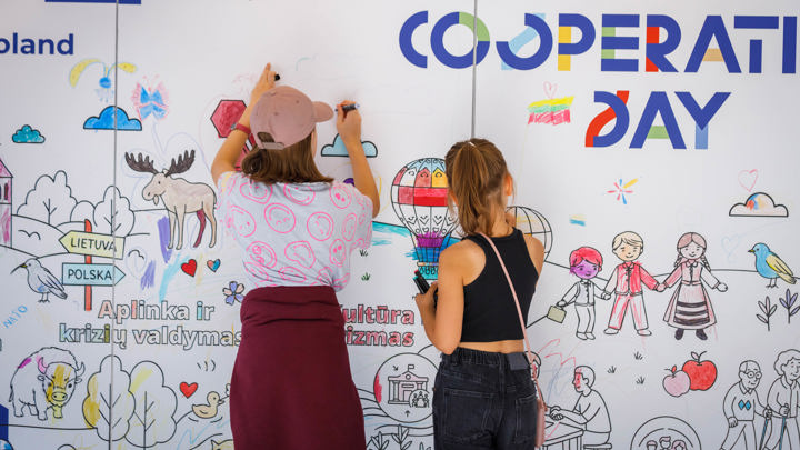 Two girls stand with their backs to the camera, coloring and drawing on a large illustrated wall decorated with characters, animals, and European Union logos promoting European Cooperation Day.