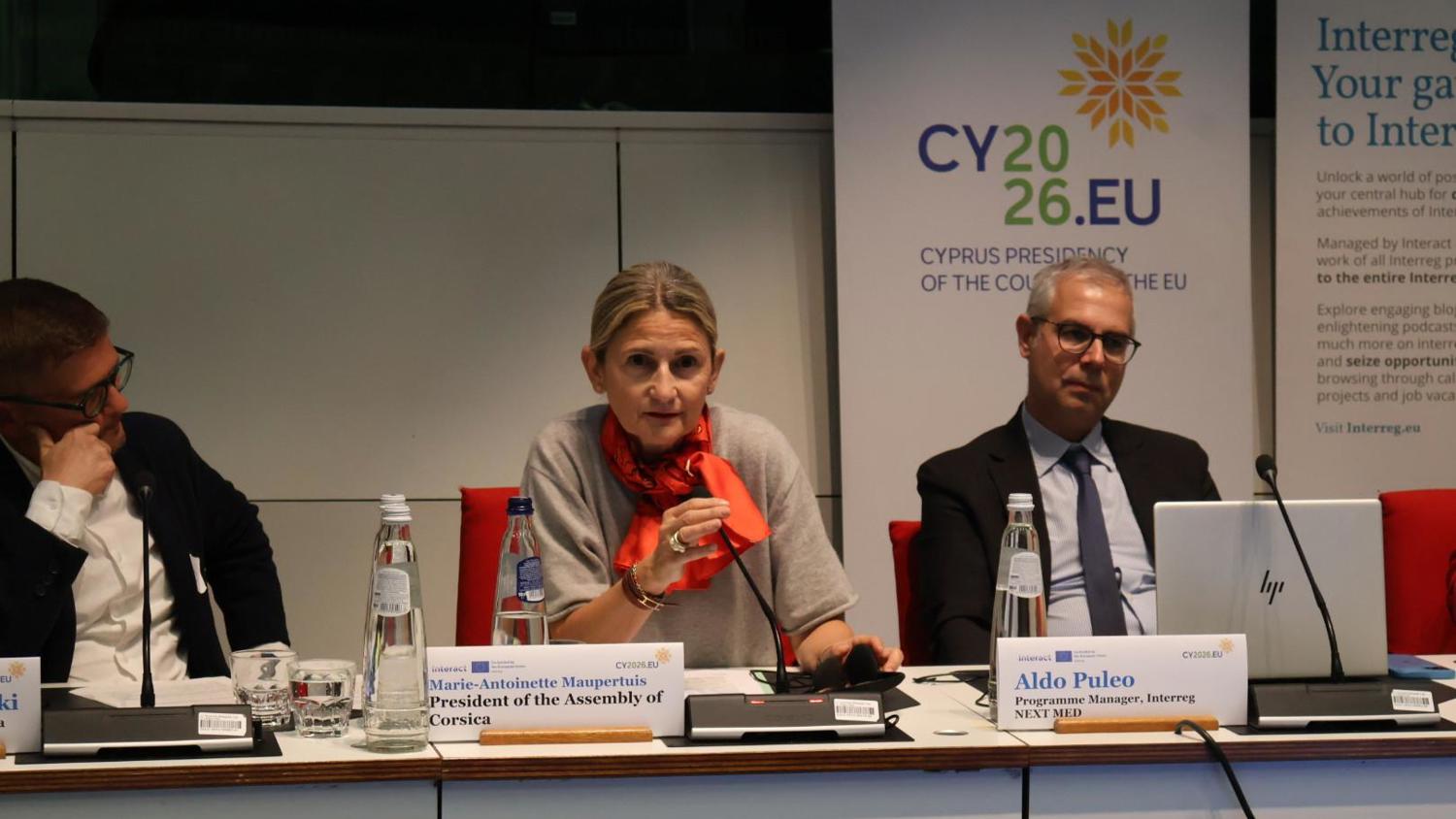 A woman speaks into a microphone at a conference table between two men. Behind them is a CY2026.EU banner and nameplates reading "Marie-Antoinette Maupertuis" and "Aldo Puleo." Bottled water and laptops are on the table.