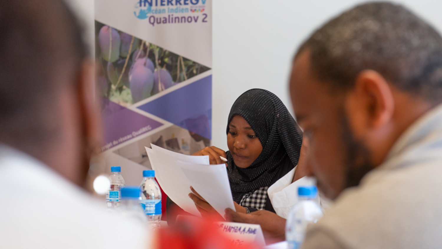 A woman wearing a hijab reads documents at a meeting. Other participants are visible, with water bottles and papers on the table. A poster with the text "INTERREG V Indian Ocean" and "Qualinnov 2" is in the background.